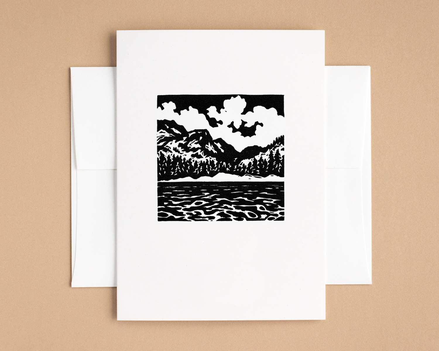 A vertical white card with a black and white depiction of a mountain lake. The card sits on top of a white envelope, which lies on top of a brown backdrop.